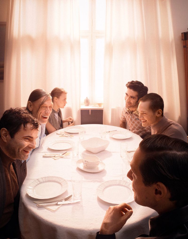 Seven people, including males and one child, are seated at a long oval table set with plates and silverware in a brightly lit room.