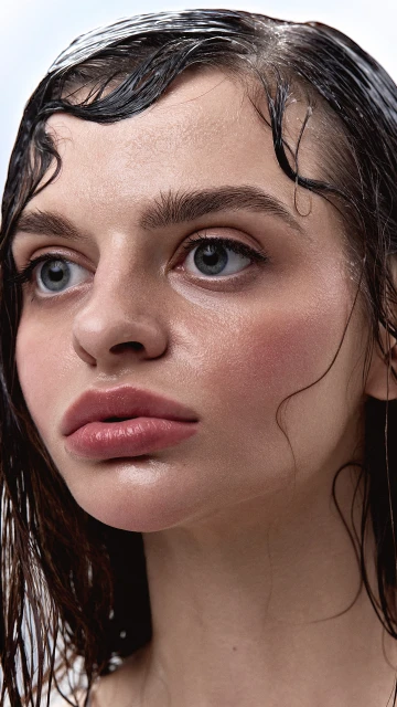 Close-up beauty portrait of a young adult female model with wet dark hair and fair skin posing in a photo studio setting with even lighting.