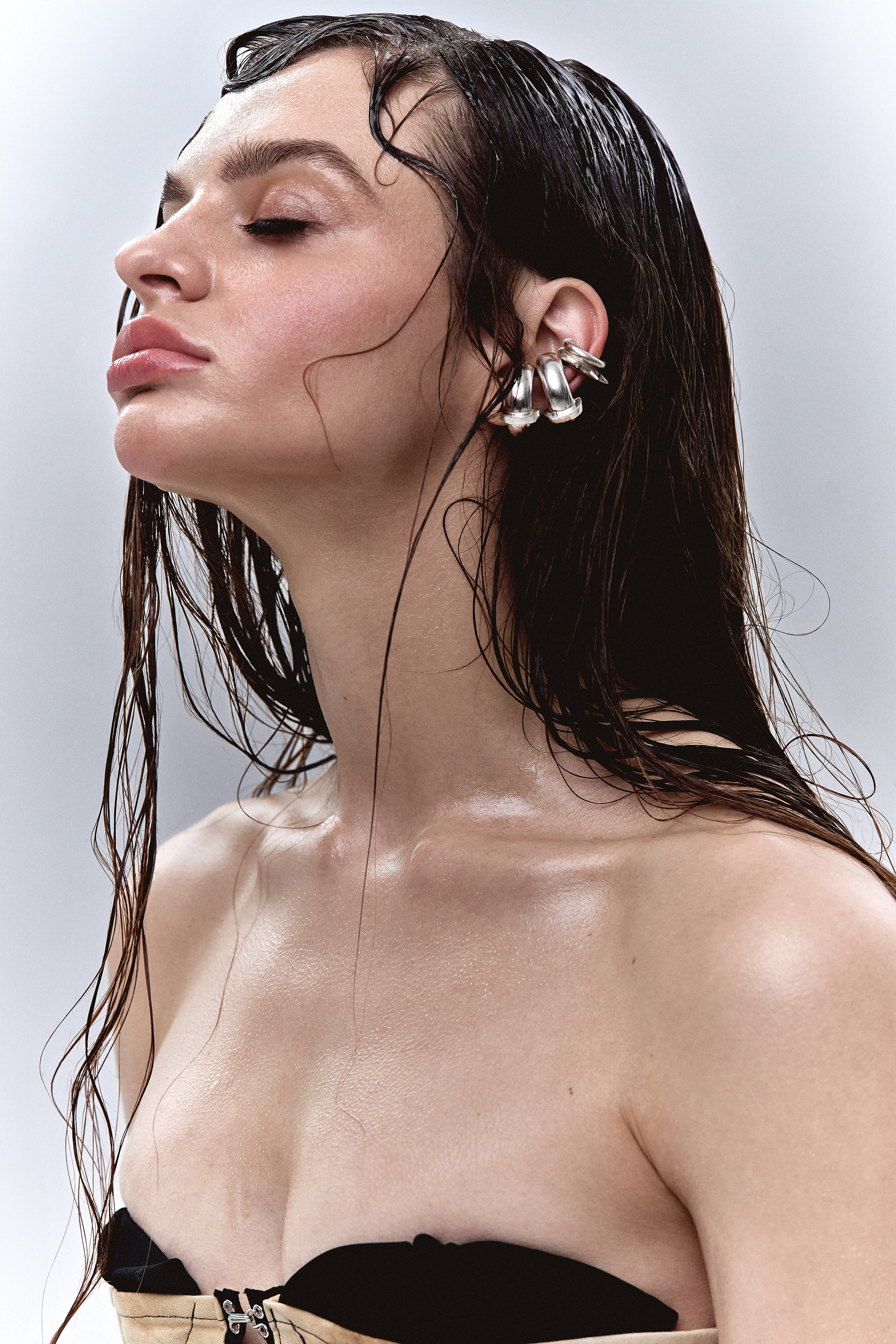Woman with wet dark hair wearing chunky silver ear cuffs tilting her head back against a neutral grey studio backdrop.