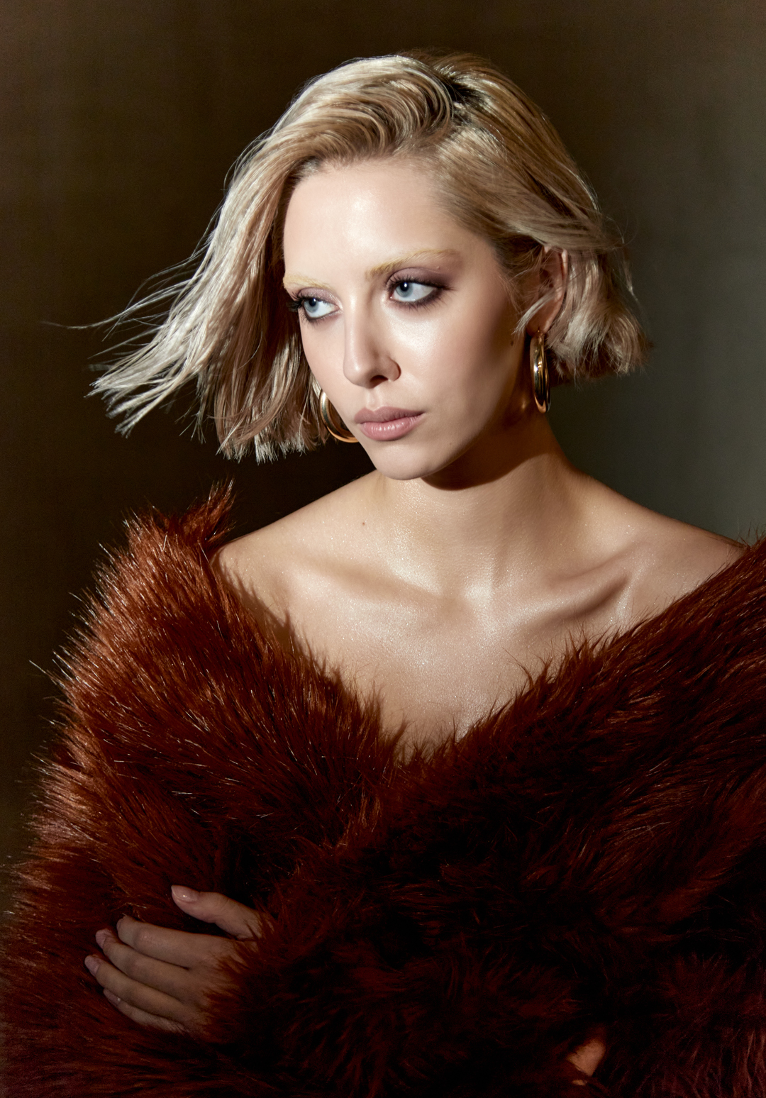 Portrait of a blonde woman with blue eyes wearing gold hoop earrings and a brown faux fur coat, posing in a studio with brown background.
