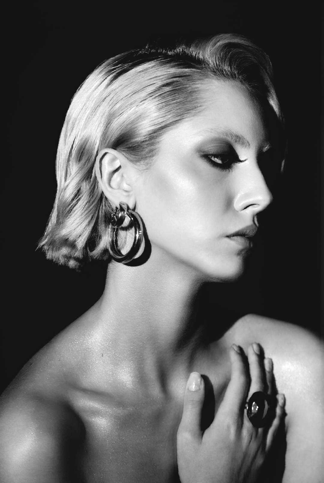 Black and white portrait of a fair-skinned woman in jewelry, side profile, with blond wavy hair, and dark eye makeup against a dark background.