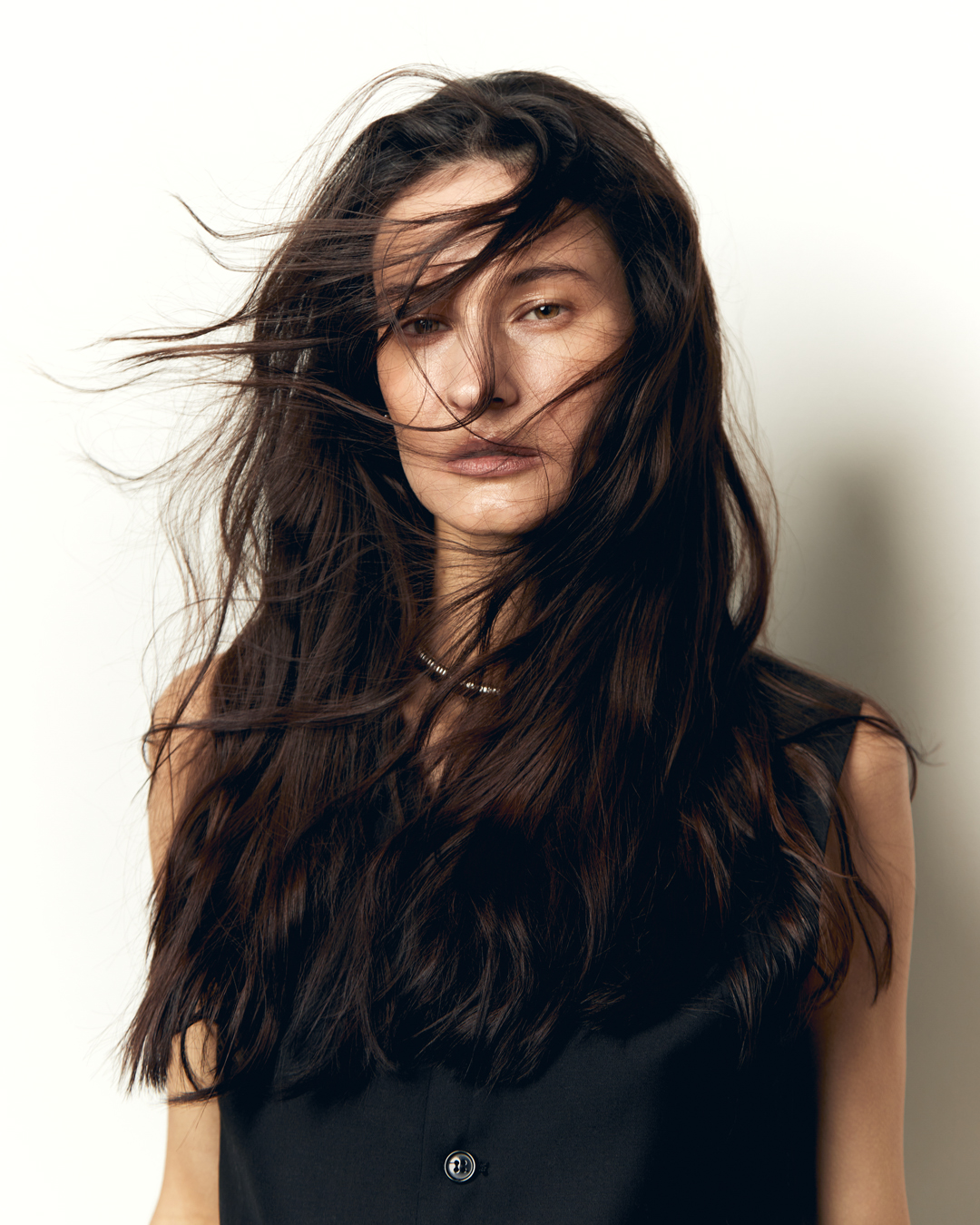 Portrait of a woman with long dark hair blowing in the wind, wearing a black top against a neutral background in an indoor setting.