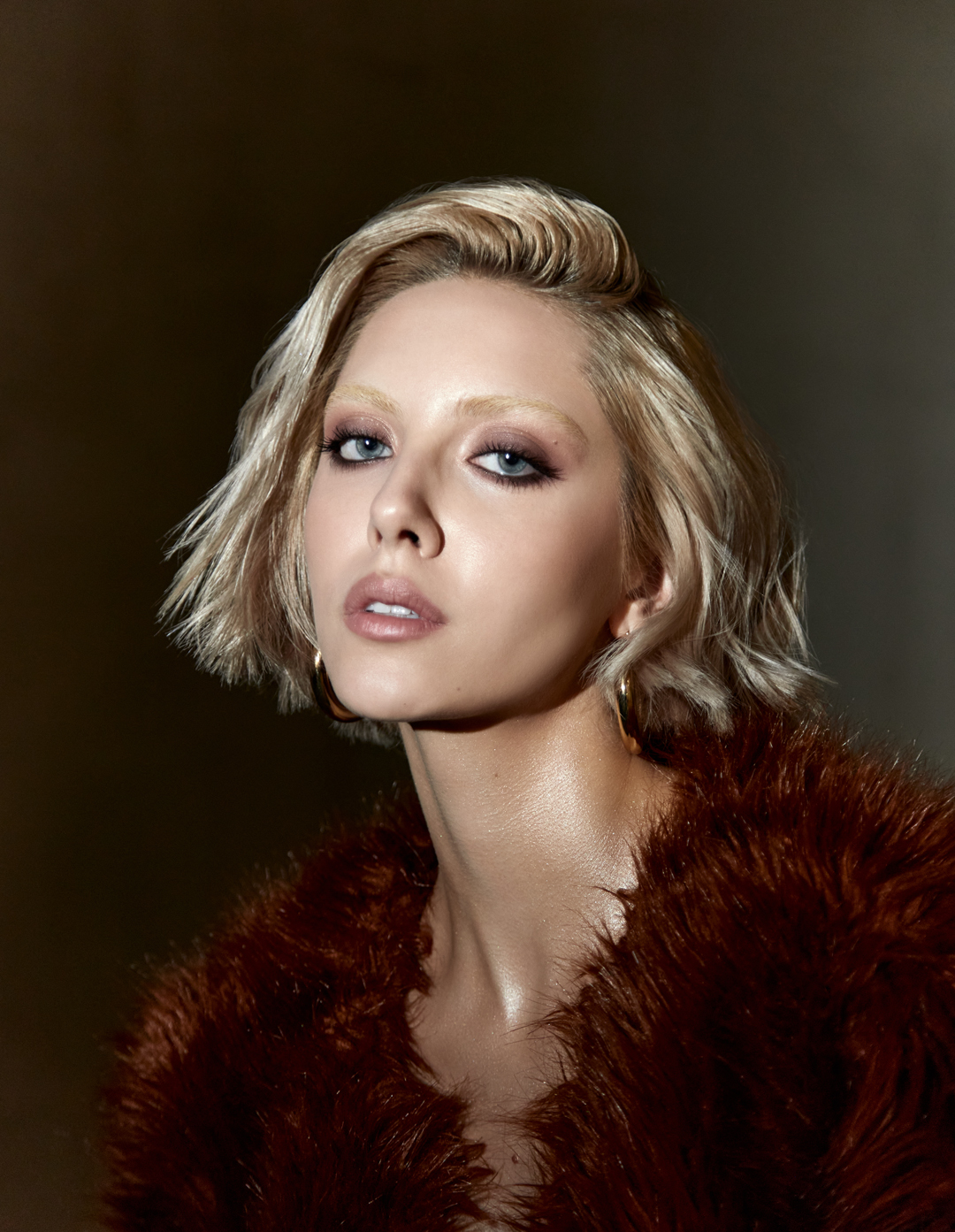 Close-up portrait of a blonde woman in a fur coat with neutral makeup against a blurred dark background.