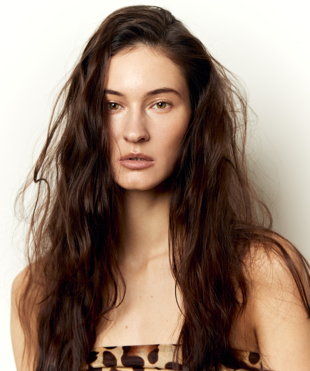 Portrait of a woman with long, wavy brown hair and fair skin, wearing a strapless leopard print top against a plain white background.