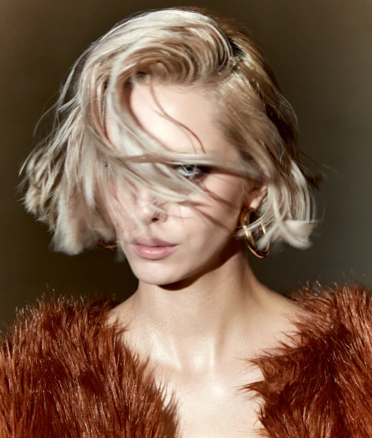 Close-up studio portrait of a blond woman with gold hoop earrings, wearing a fur jacket, in a dimly lit setting with neutral background.