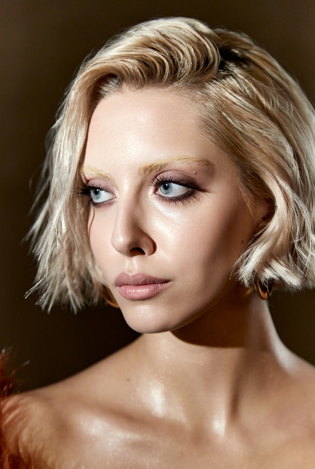 Close-up studio portrait of a young blonde woman with blue eyes, wearing gold hoop earrings and makeup, looking off to the side.