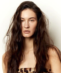 Close-up portrait of a woman with long wavy dark brown hair and natural makeup against a white studio background.