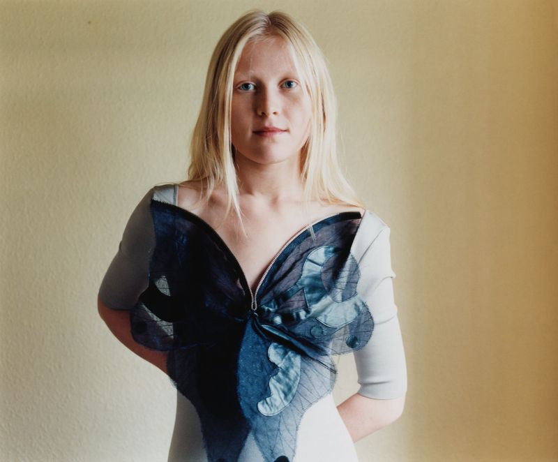 Portrait of a fair-skinned adult female with blonde hair wearing a butterfly-themed shirt against a neutral-colored wall backdrop.