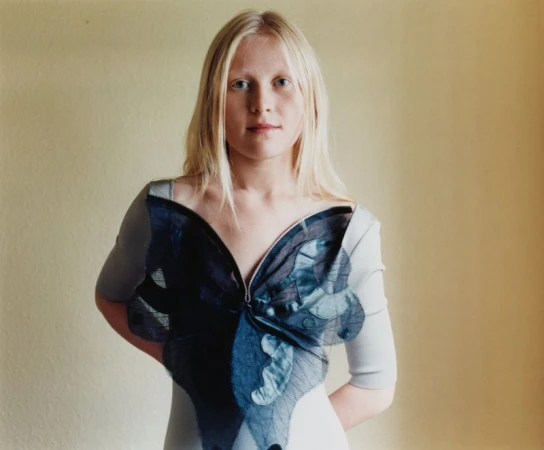 Portrait of a fair-skinned adult female with blonde hair wearing a butterfly-themed shirt against a neutral-colored wall backdrop.