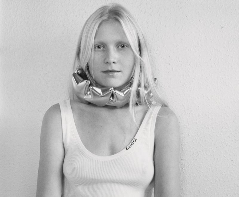 Monochrome portrait of a blonde woman wearing a metallic ruffled collar and white tank top against a textured white wall.
