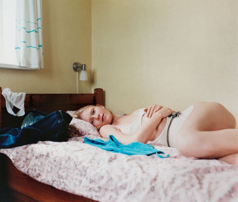 Young adult female lying on a patterned bed in a bedroom, wearing a white bodysuit and posing with her arms folded, eye-level angle.