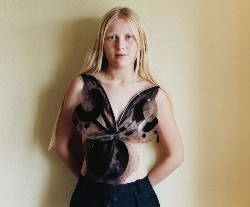 Close-up of a fair-skinned young woman with long blonde hair wearing a butterfly-shaped top in a brightly lit indoor setting.