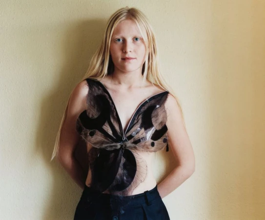 Close-up of a fair-skinned young woman with long blonde hair wearing a butterfly-shaped top in a brightly lit indoor setting.