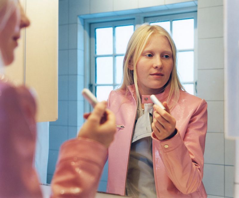 Young woman with fair hair stands in a bathroom and holds a wrapped tampon. She wears a pink jacket and is looking at a mirror.