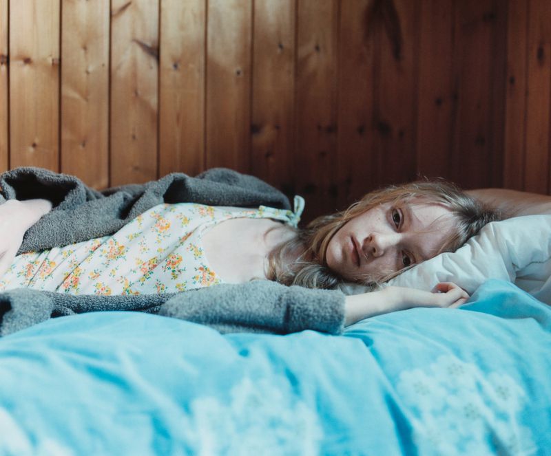A young adult woman lies in bed on a blue comforter with a pillow, covered by a gray blanket, in front of a wooden paneled wall.