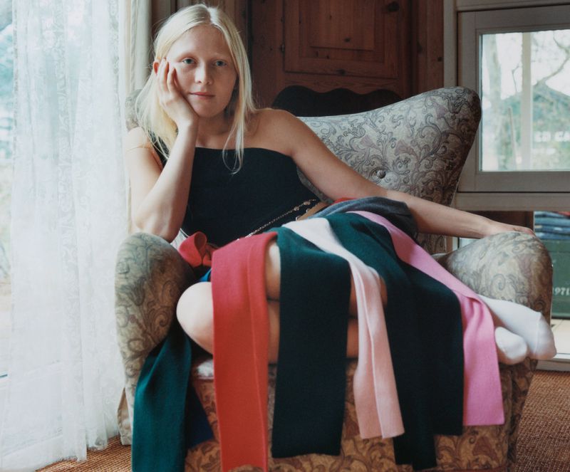 Woman with pale skin and blonde hair sits in a floral armchair surrounded by colorful scarves in a brightly lit indoor space.
