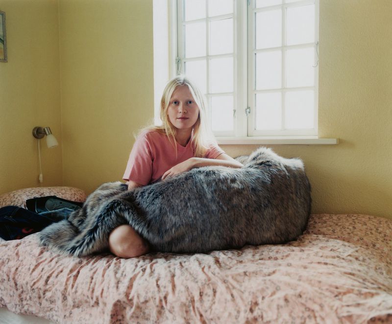 Portrait of a fair-skinned woman with blonde hair sitting on a patterned bed with a furry throw near a window in a softly lit room.