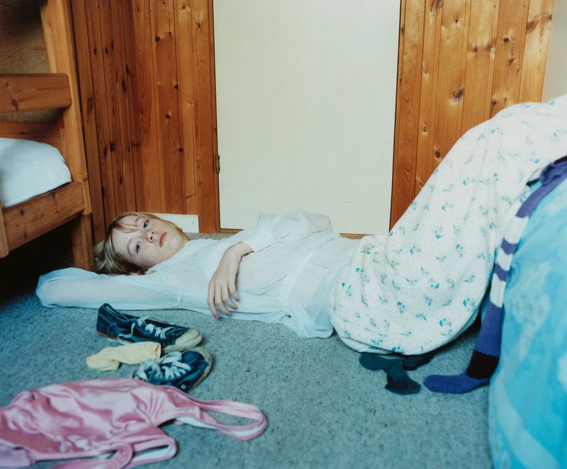 Young girl lying on the floor in pajamas in a bedroom with a bunk bed and clothing, looking at the camera with a serious expression.
