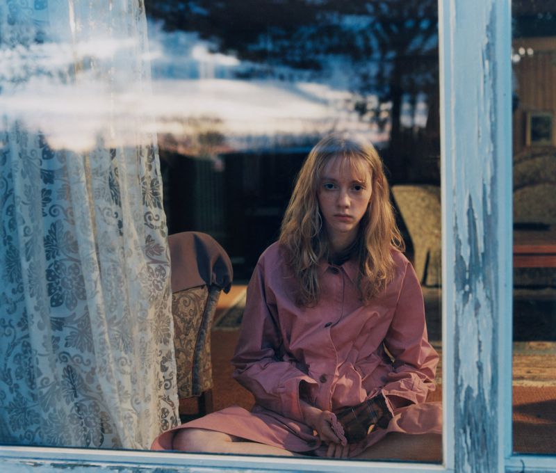 Indoor portrait of a teenage girl in a pink coat sitting near a window, reflecting a snowy landscape; creating an introspective, dreamy atmosphere.