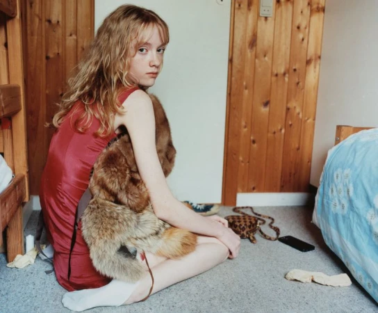 Young woman in a red dress with a fur stole sits on the floor in a simple room with wood paneling, bunkbeds, and a cell phone.