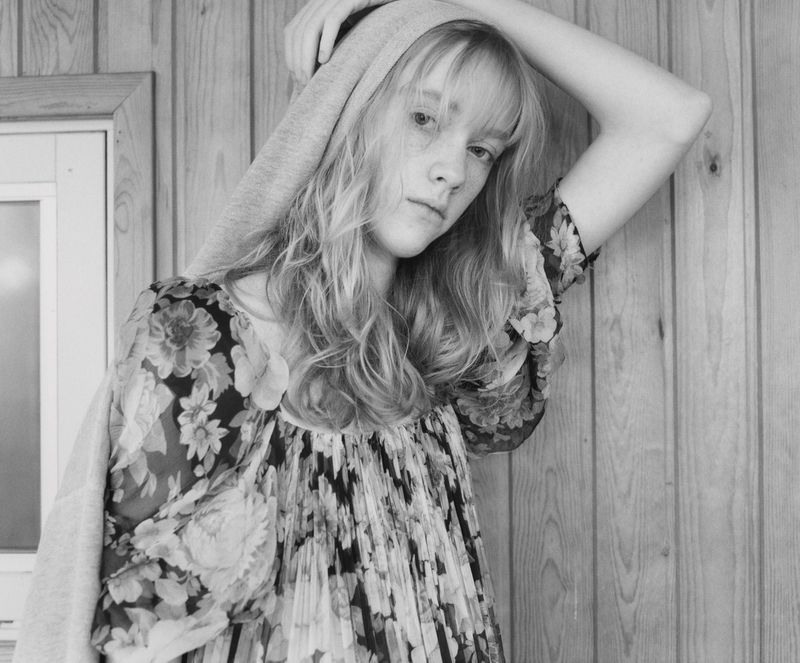 Black and white portrait of a young woman with wavy blonde hair wearing a floral dress and light grey hoodie, posing indoors.
