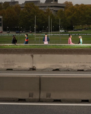 Group of people walking along an elevated concrete barrier next to a highway, with a green field and buildings in the background.