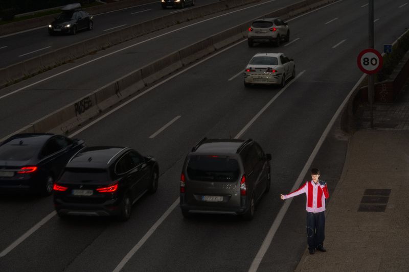 Person in red and white jacket stands on roadside near highway with several cars, and speed limit sign indicating 80 kilometers per hour.