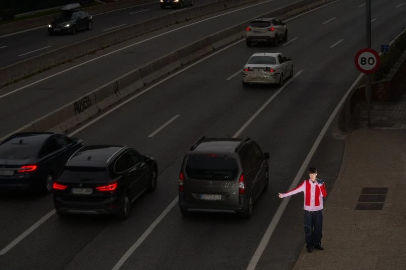 Person in red and white jacket stands on roadside near highway with several cars, and speed limit sign indicating 80 kilometers per hour.