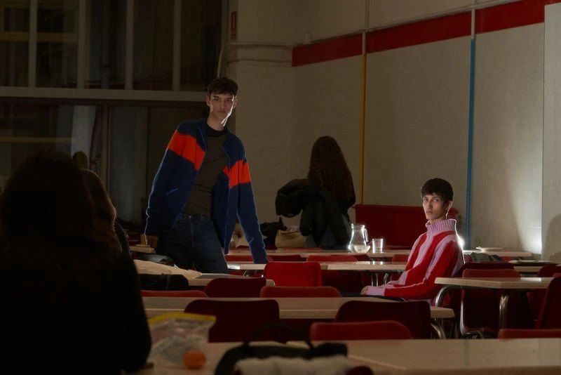 Two young adult men in casual fashion stand in a dimly lit room with tables, chairs, and other people, exuding a stylish and relaxed vibe.