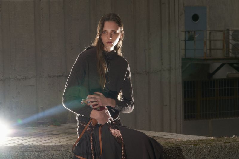 Adult woman in casual wear with long brown hair poses in an urban outdoor setting with concrete walls under bright, diffused lighting.