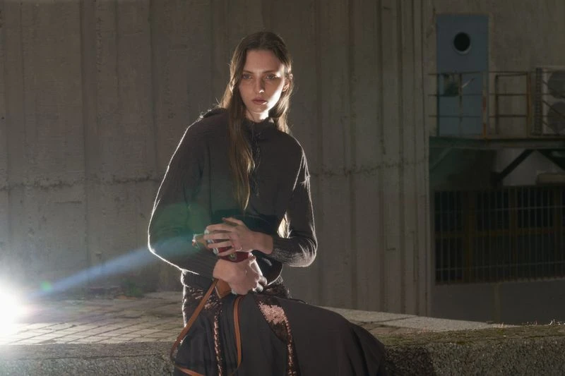 Adult woman in casual wear with long brown hair poses in an urban outdoor setting with concrete walls under bright, diffused lighting.
