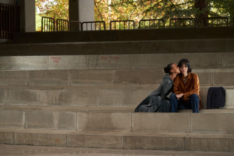 Two women sit on concrete steps outdoors in a public space, one kissing the other's cheek. Autumn leaves on trees in the background.