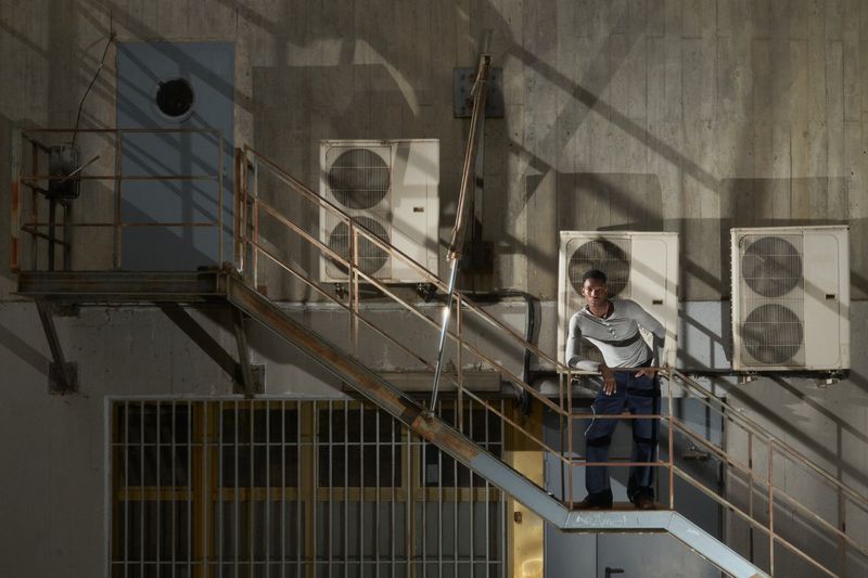 A man stands on metal stairs against a concrete building, featuring AC units, a barred window, and geometric shadow patterns.