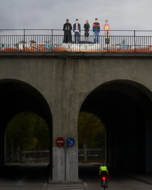 Group of five young adults stand on a bridge above a road with a cyclist, featuring urban architecture and overcast sky.