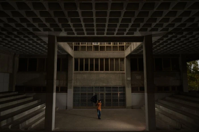 Figure standing in a courtyard framed by brutalist concrete columns and coffered ceiling for Eternal Chase