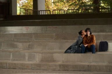 Two models sitting on wide concrete steps outdoors, one kissing the cheek of the other in an editorial campaign photograph.