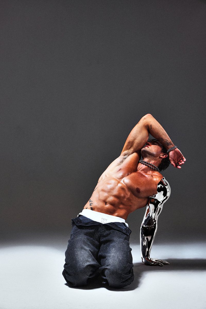 An athletic, muscular adult male with a metallic arm poses on his knees against a gray backdrop, showcasing body art and jewelry.