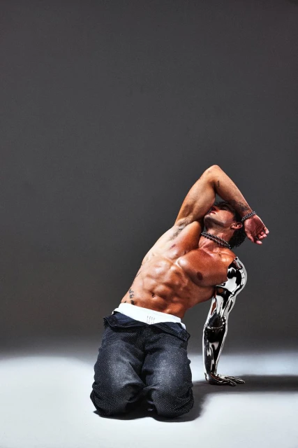 An athletic, muscular adult male with a metallic arm poses on his knees against a gray backdrop, showcasing body art and jewelry.