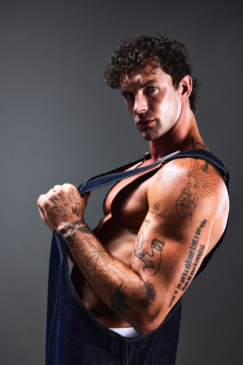 Muscular man poses shirtless with curly hair and tattoos in a studio, wearing denim overalls against a grey background.