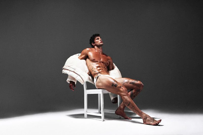 Athletic man with tattoos sits on a white chair, wearing a white jacket and swimsuit bottoms in a light studio against a gray background.