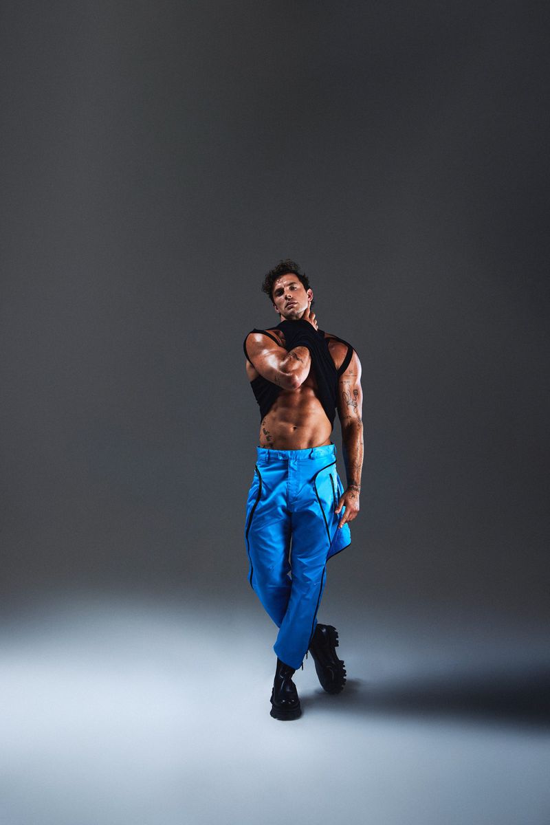 Athletic man poses in black top and blue pants in a studio. He stands with his arms crossed, showcasing abs, and tattoos on his arms.