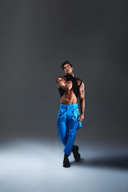 Athletic man poses in black top and blue pants in a studio. He stands with his arms crossed, showcasing abs, and tattoos on his arms.