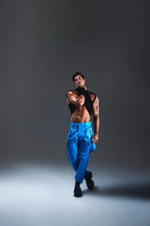 Athletic man poses in black top and blue pants in a studio. He stands with his arms crossed, showcasing abs, and tattoos on his arms.