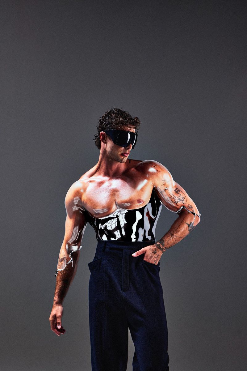 A muscular man poses in black pants with white body paint and dark sunglasses, set against a neutral gray backdrop in a studio setting.