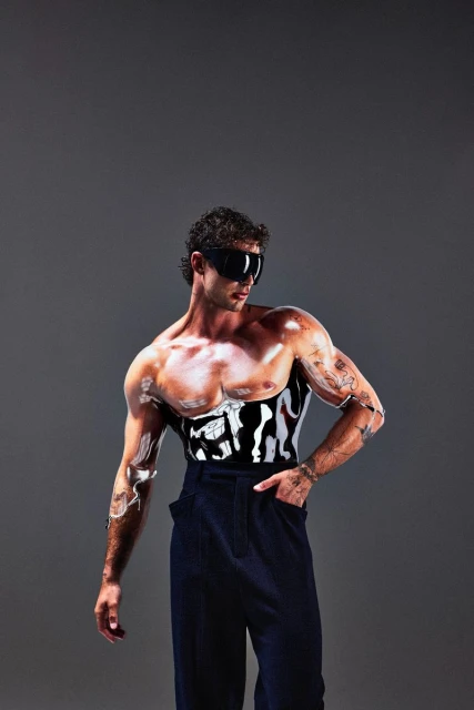 A muscular man poses in black pants with white body paint and dark sunglasses, set against a neutral gray backdrop in a studio setting.