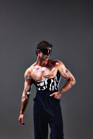 A muscular man poses in black pants with white body paint and dark sunglasses, set against a neutral gray backdrop in a studio setting.