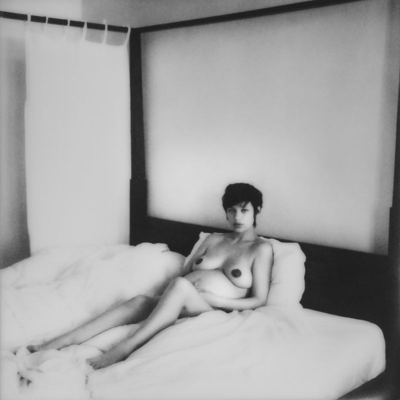 A nude, pregnant adult woman with short dark hair lies on a bed with white sheets in an indoor setting, looking directly at the camera.