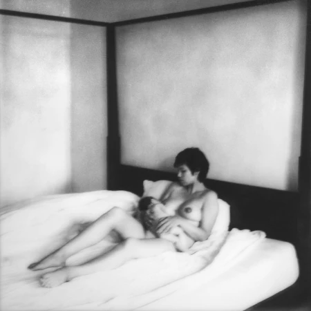 Monochromatic portrait of an adult female breastfeeding her baby in a bedroom, featuring a four-poster bed with white linens.