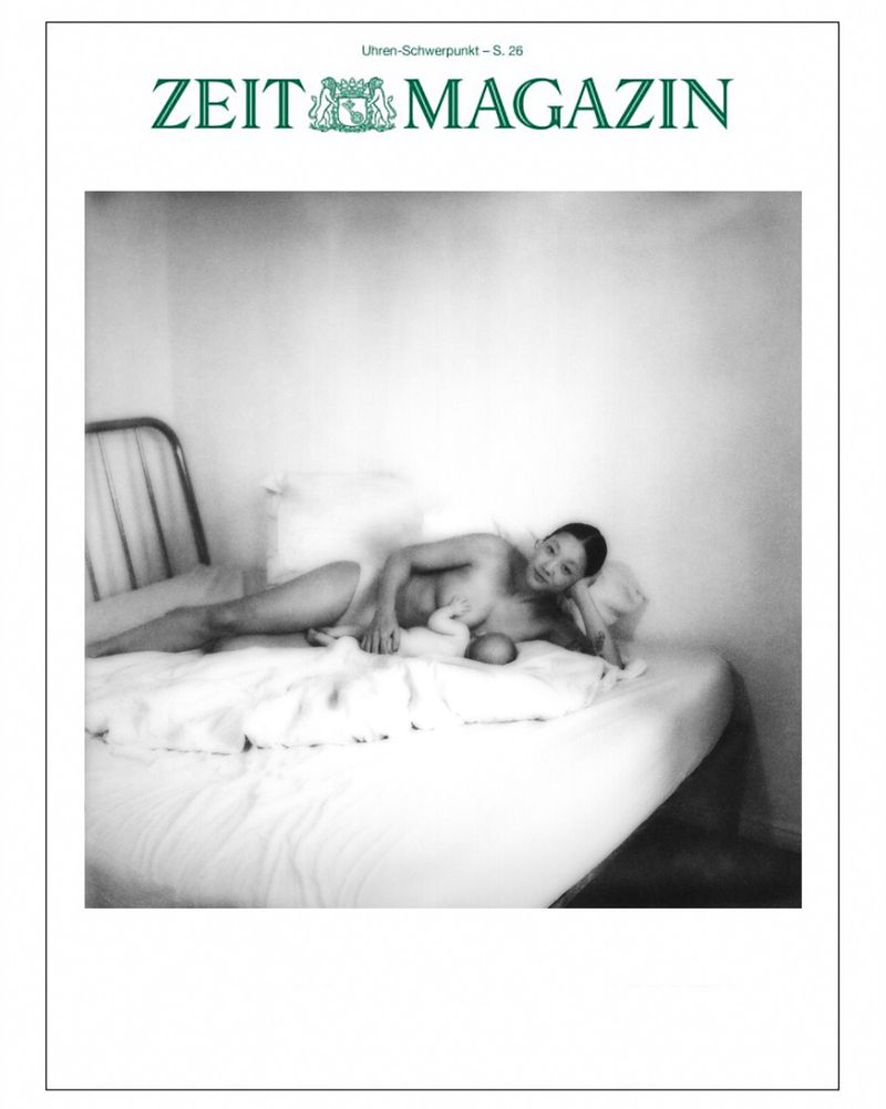 Black-and-white magazine cover featuring a nude woman lying on a bed with a baby, minimal interior setting, and natural lighting.