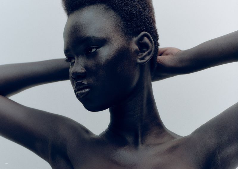 Close-up portrait of a Black woman with short hair, her hands behind her head, against a white background. Focus on the face.